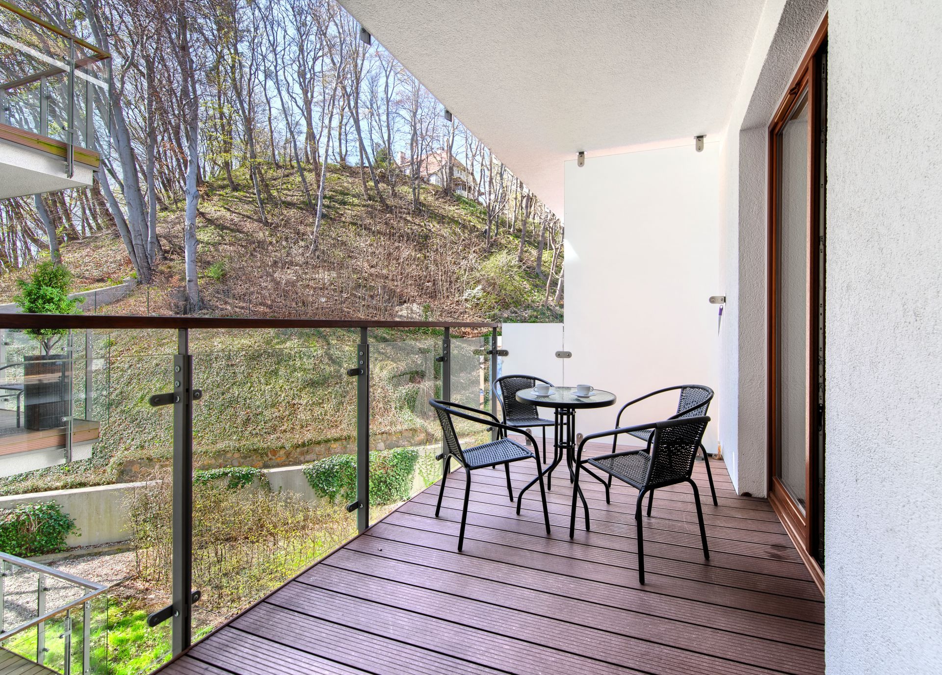 Zadaszony balkon z drewnianą podłogą, szklaną balustradą i widokiem na zalesione wzgórze. Cztery czarne krzesła z plecionki i okrągły stolik z dwiema filiżankami.