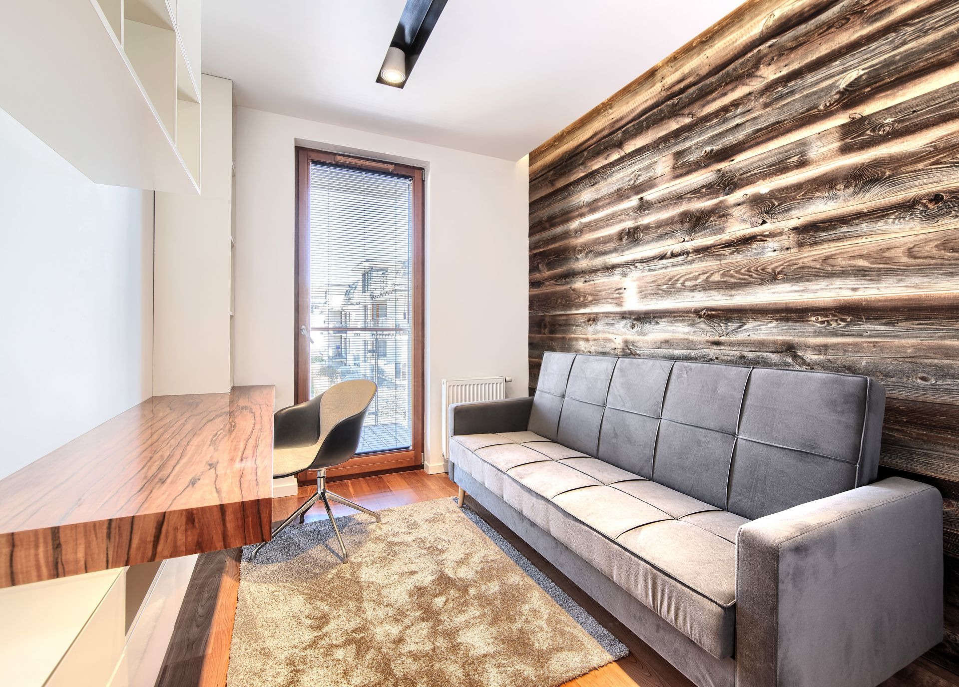 Nowoczesny pokój z biurkiem z drewnianym blatem, szarą sofą na tle ściany z desek, dywanem i oknem z żaluzjami.