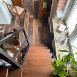 Widok z góry na drewniane schody z metalową balustradą prowadzące w dół, z widocznym fragmentem drewnianej podłogi i mebli w tle oraz rośliną doniczkową przy oknie.