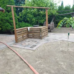 Ogród z kostki brukowej z drewnianym barem zbudowanym z palet, oświetlonym girlandą żarówek, w tle dekoracja z drewnianych kół.