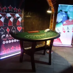 Stół do blackjacka z żetonami i kartami, otoczony podświetlanymi bannerami kasynowymi w ceglanym pomieszczeniu.