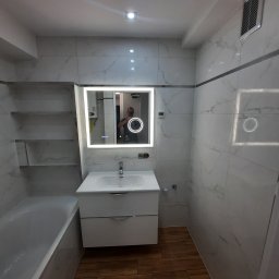 Usługi Ogólnobudowlane Paweł Krawczyk - Nowoczesna łazienka z białymi marmurkowymi płytkami, drewnianą podłogą, podświetlanym lustrem i wanną z parawanem.