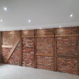 Usługi Ogólnobudowlane Paweł Krawczyk - Wykończona ściana z czerwonej cegły z drewnianymi belkami, oświetlona punktowymi lampami sufitowymi, na podłodze jasne płytki