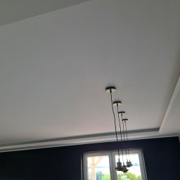 Usługi Ogólnobudowlane Paweł Krawczyk - Szary sufit podwieszany z dekoracyjną listwą i czterema wiszącymi lampami bez kloszy, widok na okno z zielenią w tle.