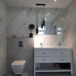 Nowoczesna łazienka z marmurowymi płytkami, wiszącą toaletą, białą szafką pod umywalką i potrójną lampą wiszącą z cylindrycznymi kloszami.