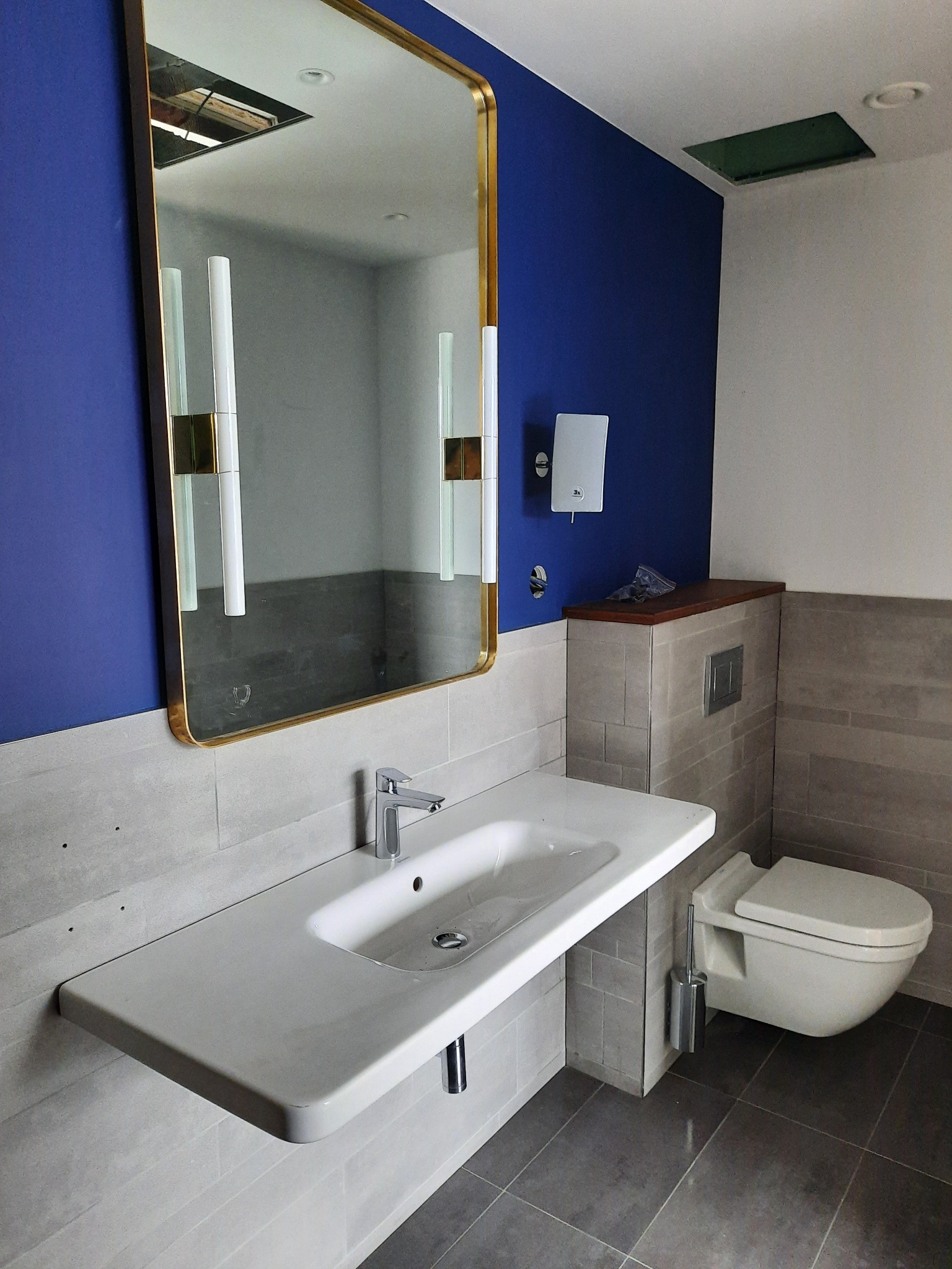 Nowoczesna łazienka z umywalką nablatową, podwieszaną toaletą i lustrem z oświetleniem, ściany wykończone w odcieniach szarości i granatu, widoczna otwarta przestrzeń w suficie.