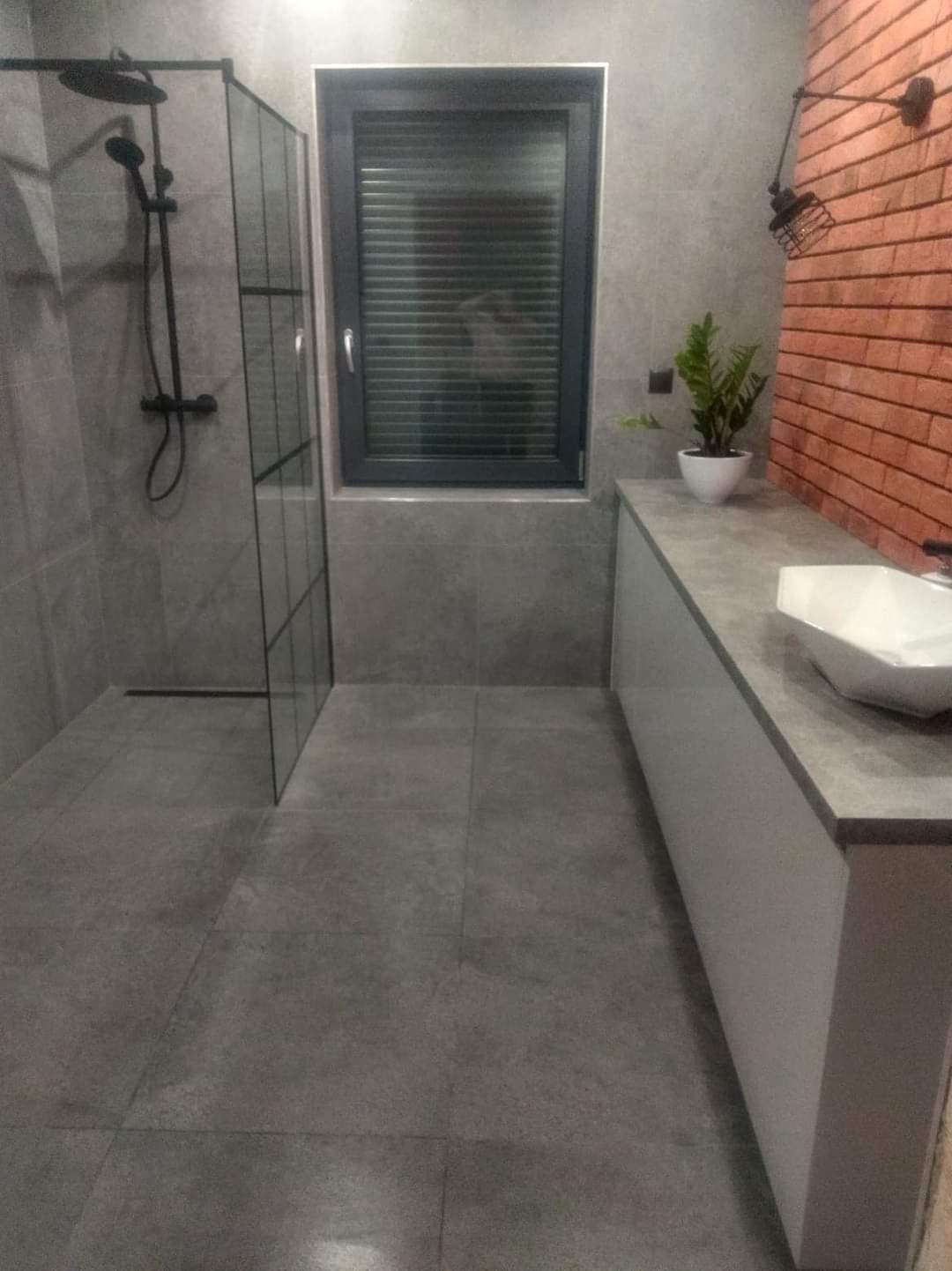 Nowoczesna łazienka z szarą glazurą, kabiną prysznicową typu walk-in z czarnymi profilami, białą szafką pod umywalką i akcentem ściany z cegły.