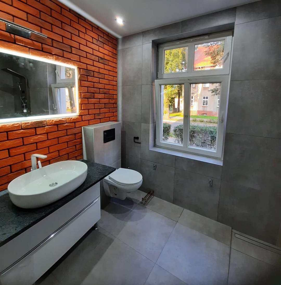 Nowoczesna łazienka z ceglaną ścianą, owalną umywalką na szafce i szarą glazurą, widok na zewnątrz przez okno.