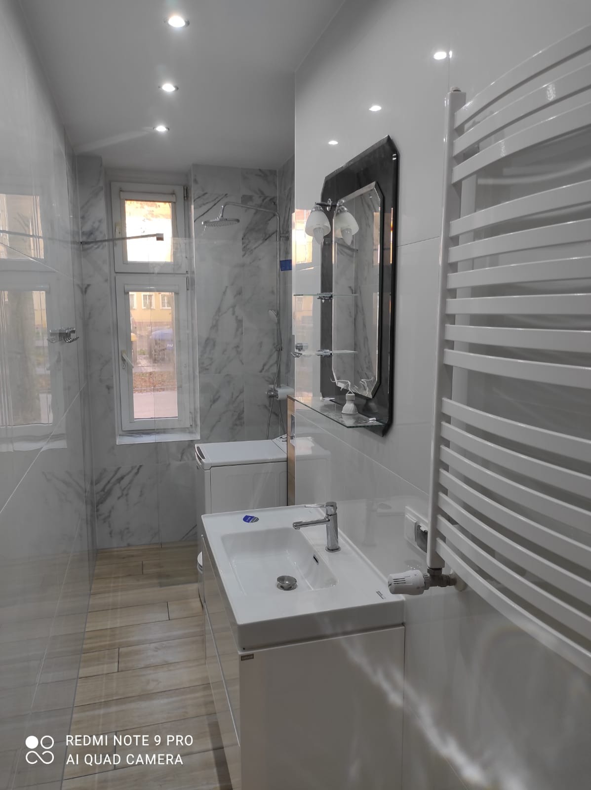 Wykończona łazienka z marmurowymi płytkami, białą szafką z umywalką, grzejnikiem drabinkowym i lustrem z oświetleniem, widoczna pralka i okno.