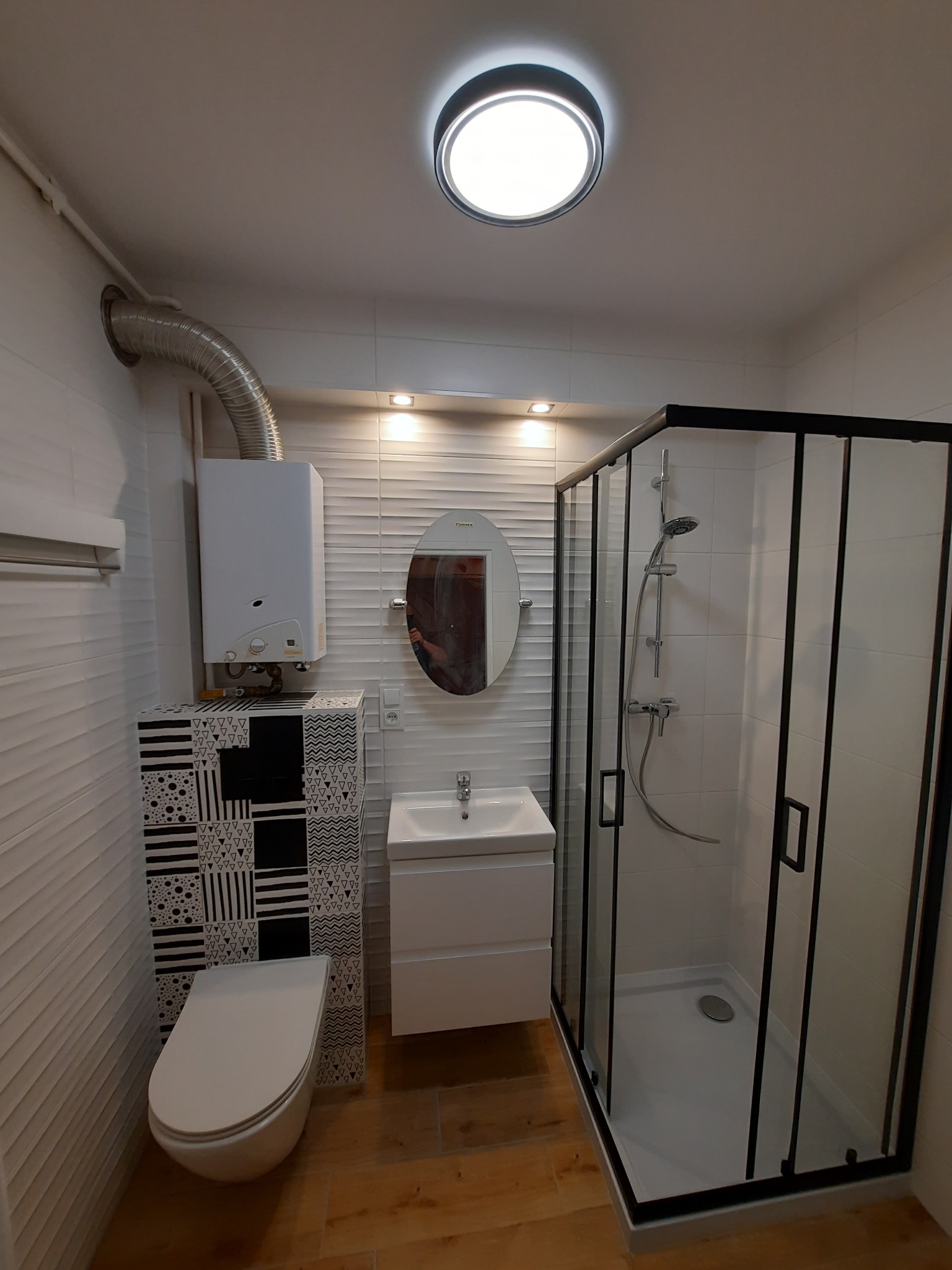 Nowoczesna łazienka z kabiną prysznicową z czarnymi ramami, podwieszaną toaletą z dekoracyjną obudową, białą umywalką z szafką i owalnym lustrem, oświetlona lampą sufitową i listwą LED.