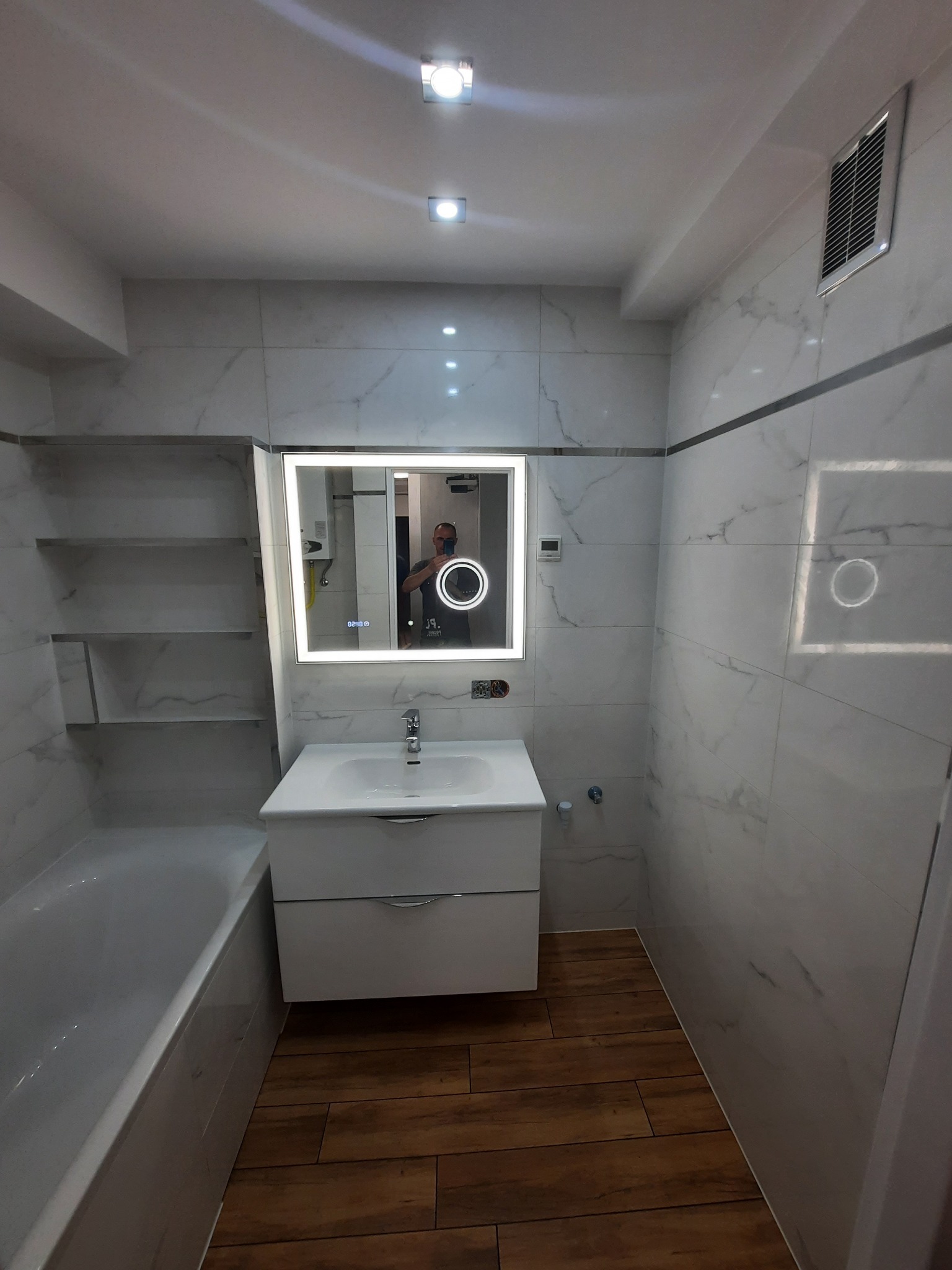 Nowoczesna łazienka z białymi marmurkowymi płytkami, drewnianą podłogą, podświetlanym lustrem i wanną z parawanem.