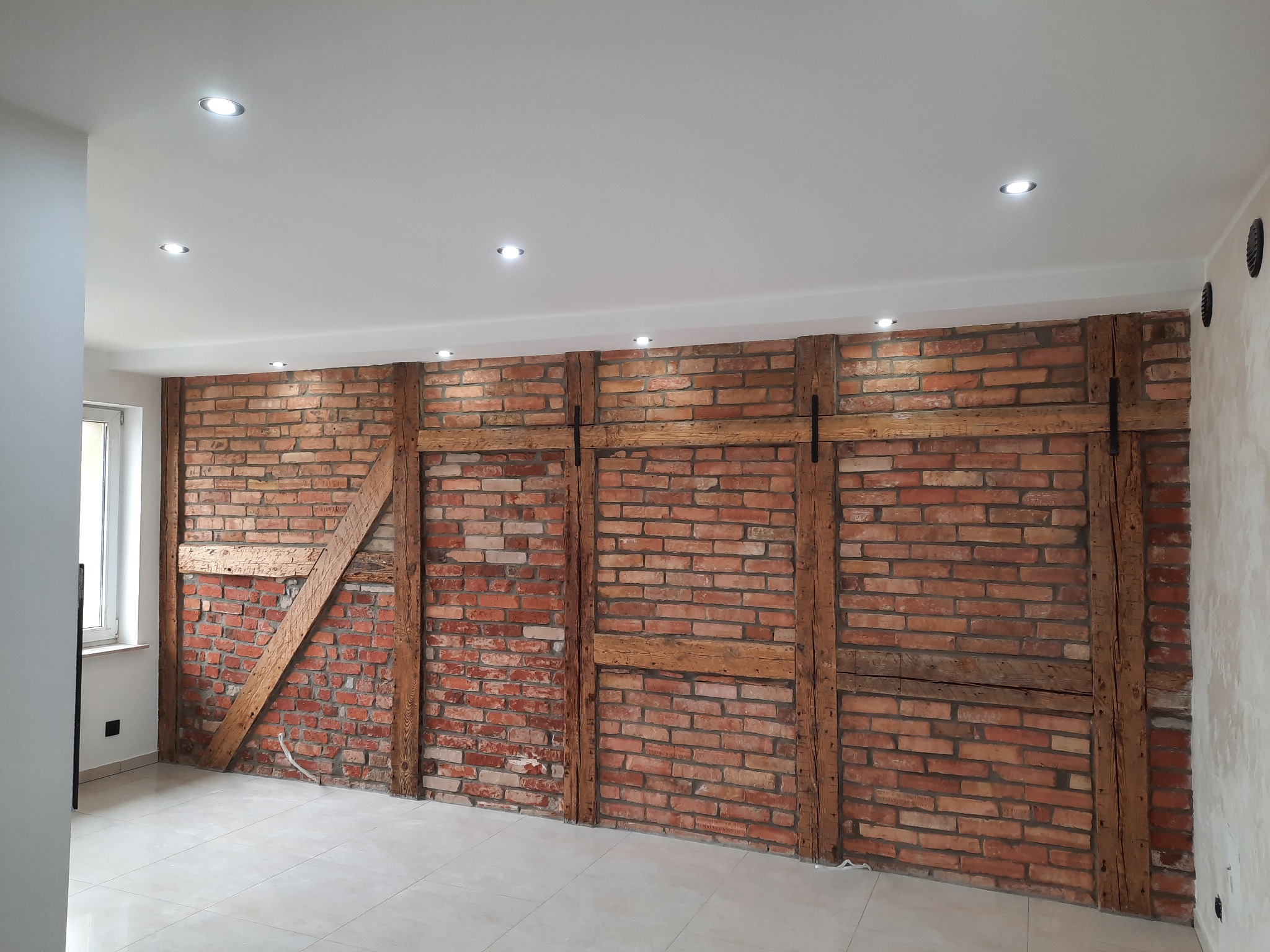 Wykończona ściana z czerwonej cegły z drewnianymi belkami, oświetlona punktowymi lampami sufitowymi, na podłodze jasne płytki