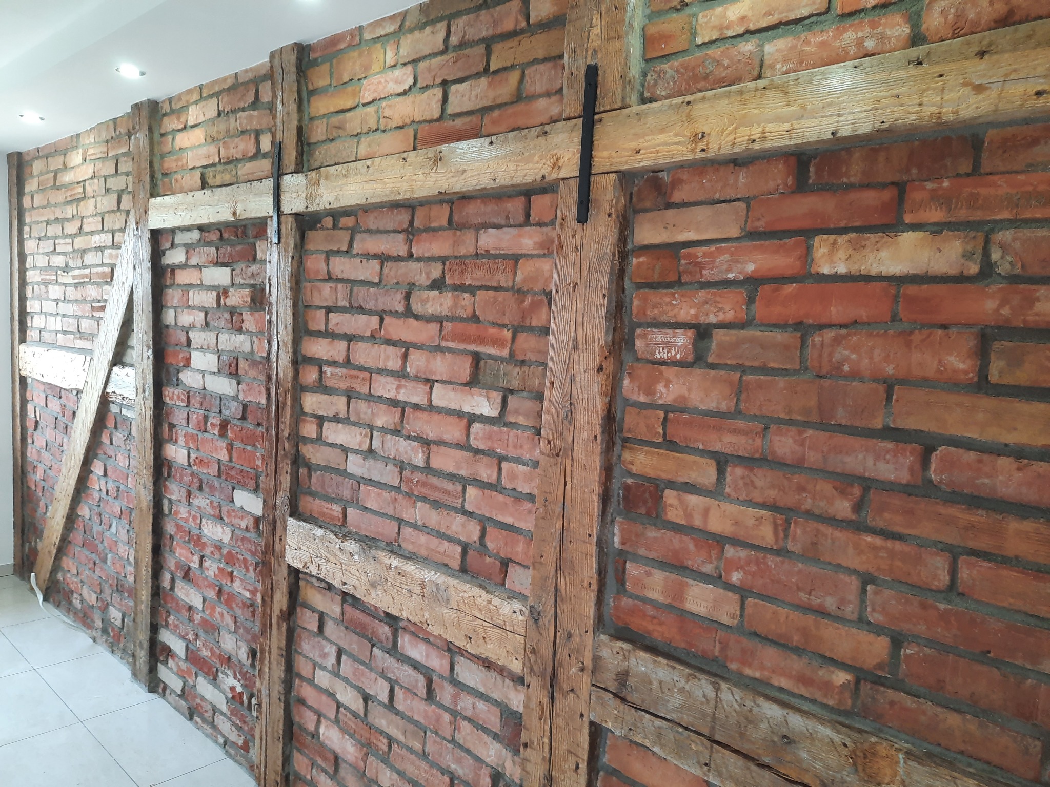 Ceglana ściana wewnętrzna z drewnianą konstrukcją szkieletową, widoczne ślady napraw i renowacji, oświetlenie sufitowe.