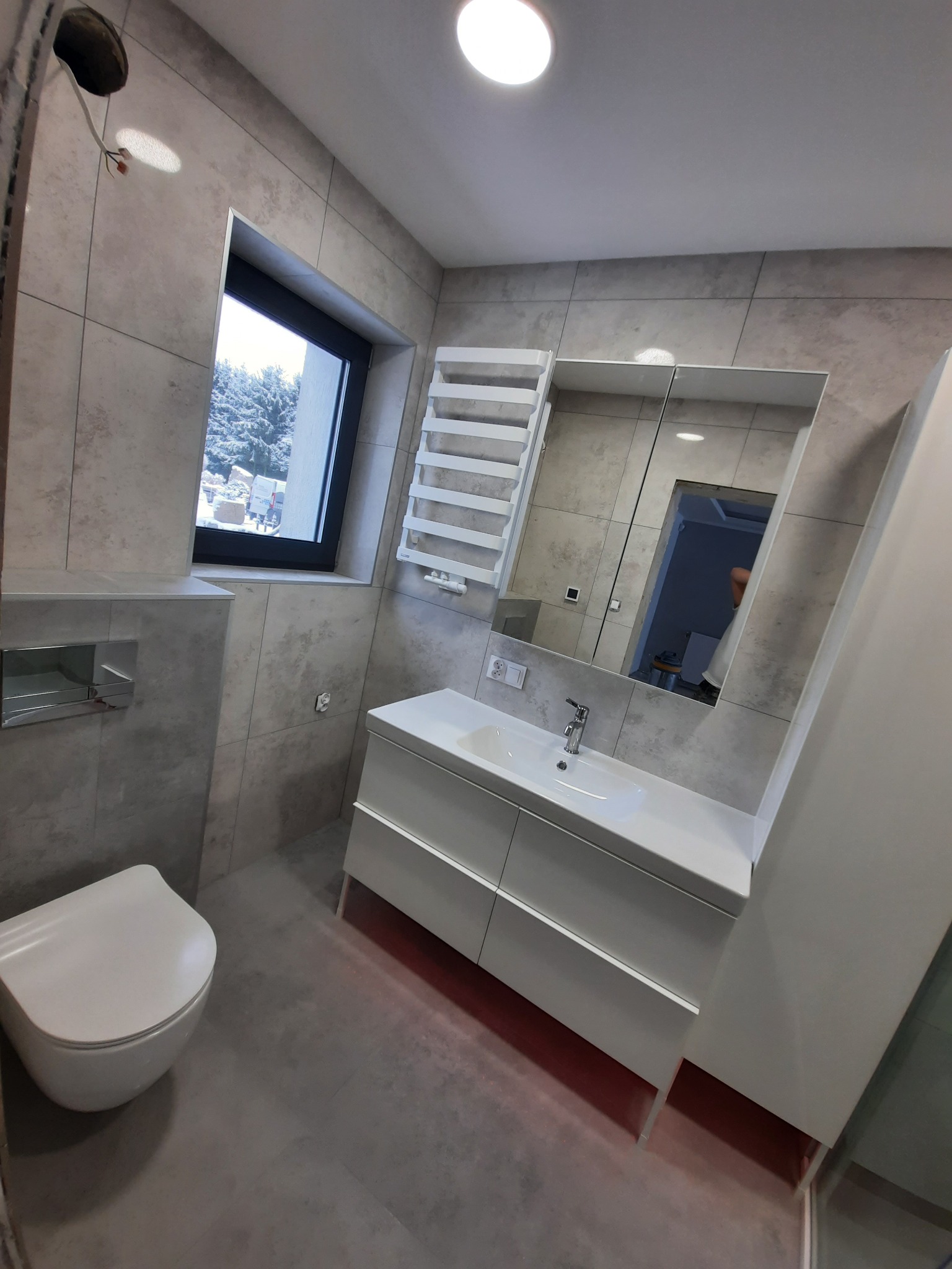 Nowoczesna łazienka z szarymi płytkami, białą umywalką z szafką, toaletą i grzejnikiem drabinkowym; widok przez okno na ośnieżone drzewa.