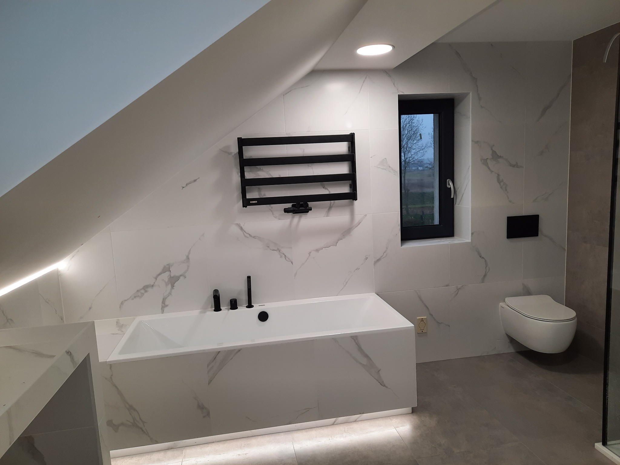 Nowoczesna łazienka na poddaszu z białą wanną obudowaną płytkami imitującymi marmur, czarną armaturą i grzejnikiem drabinkowym, oraz oknem z widokiem na zewnątrz.