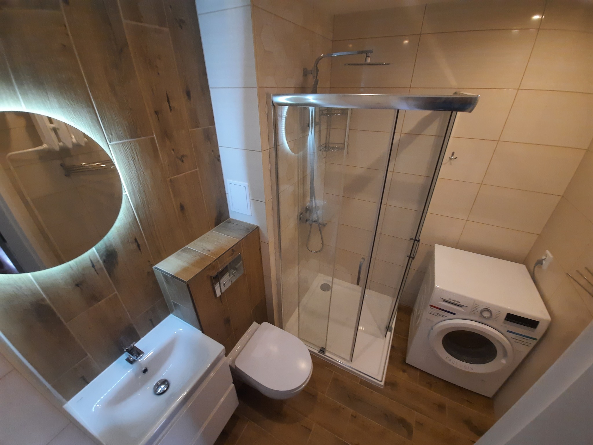 Nowoczesna łazienka z drewnopodobnymi płytkami, oświetlonym lustrem, kabiną prysznicową, toaletą podwieszaną i pralką Bosch.