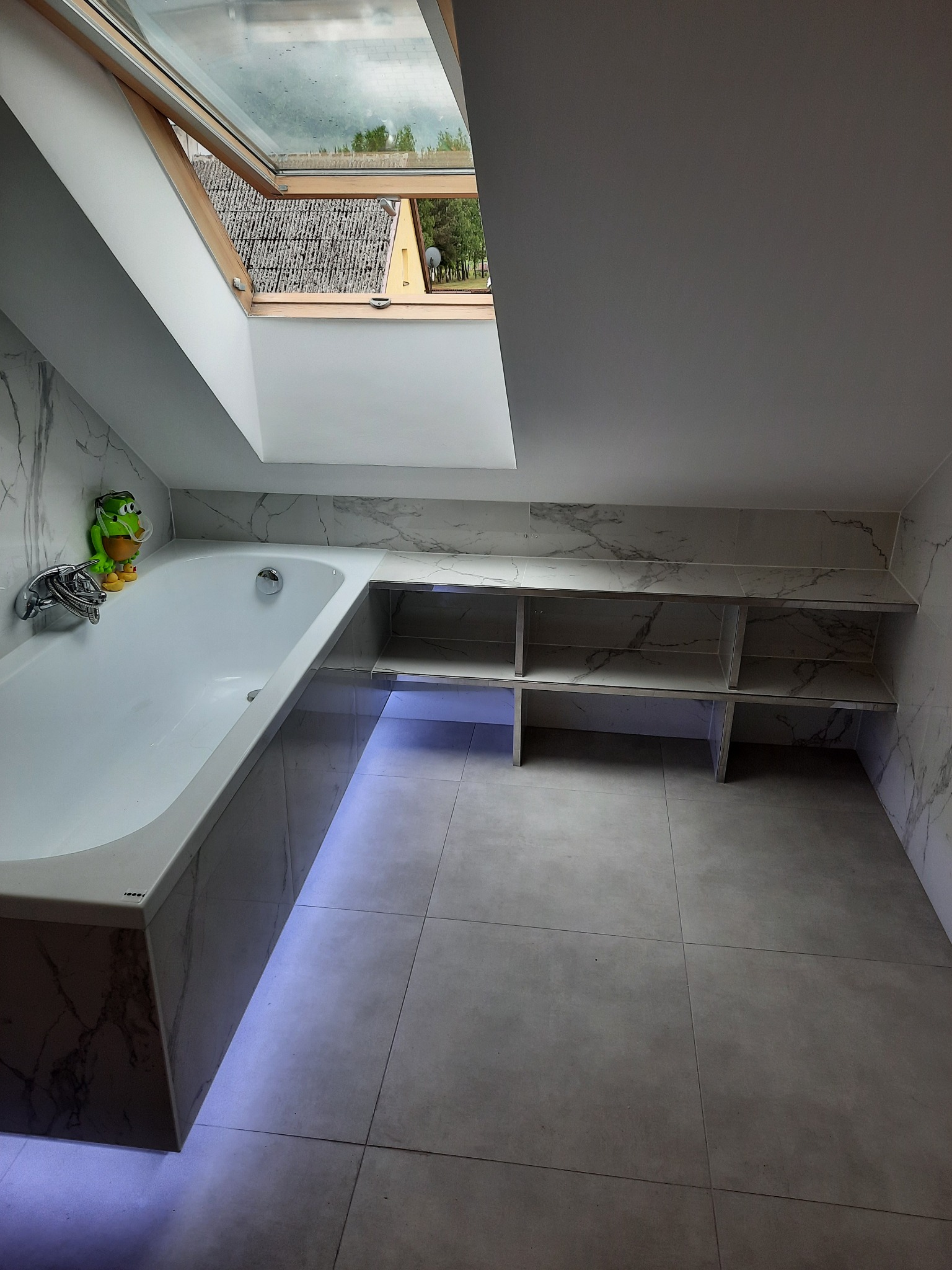 Nowoczesna łazienka na poddaszu z wanną obudowaną marmurowymi płytkami, podświetleniem LED u dołu, półkami w ścianie wyłożonymi tymi samymi płytkami oraz oknem dachowym z widokiem na dom i zieleń.