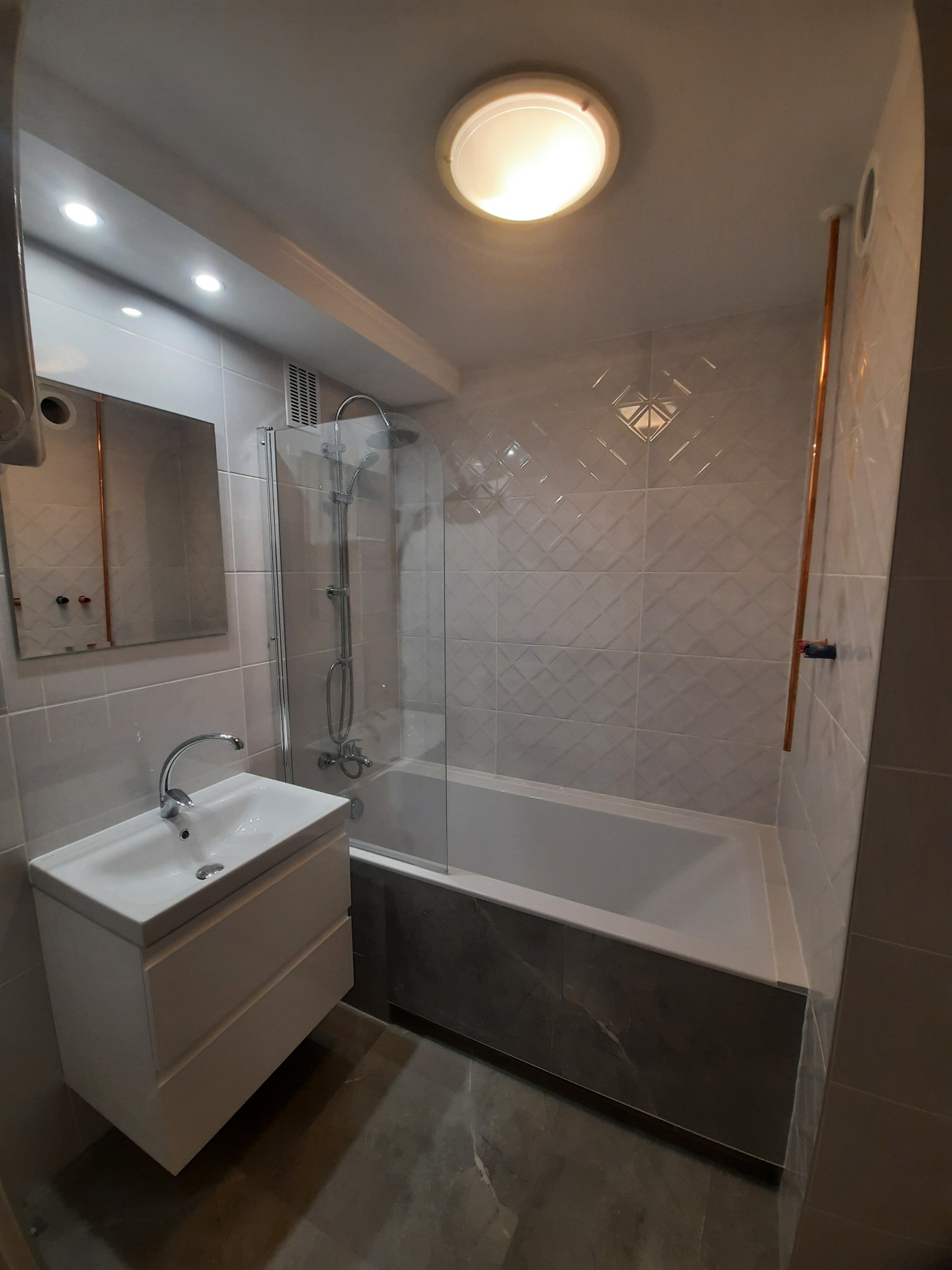 Wykończona łazienka w szaro-białej kolorystyce z wanną za szklanym parawanem, umywalką z szafką i dekoracyjnymi płytkami na ścianie.