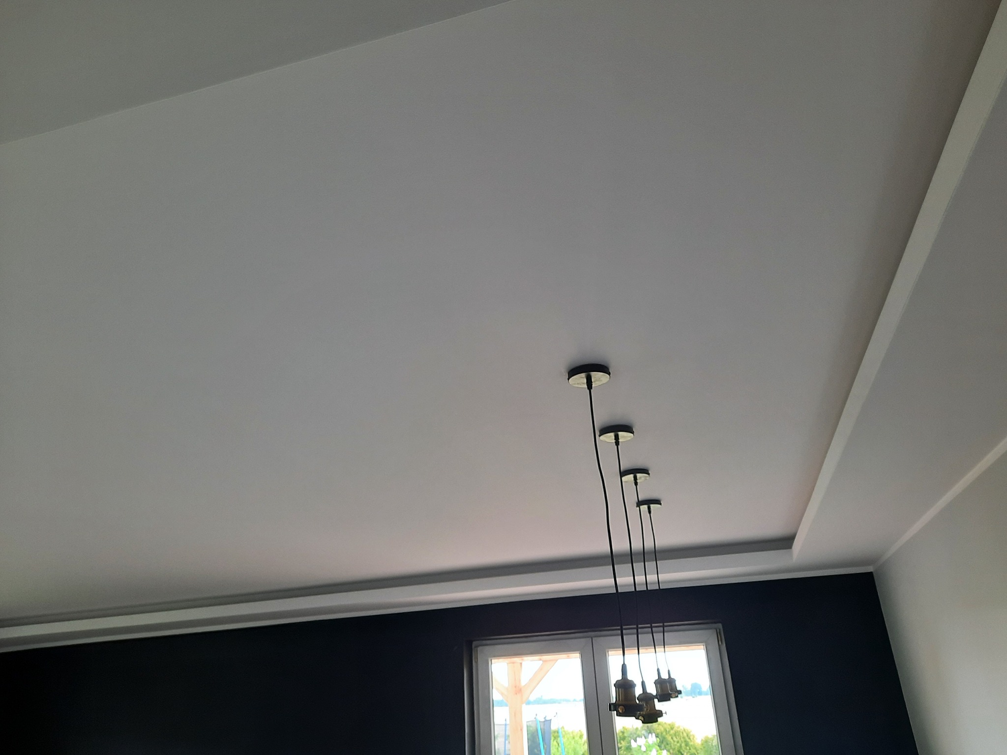 Szary sufit podwieszany z dekoracyjną listwą i czterema wiszącymi lampami bez kloszy, widok na okno z zielenią w tle.