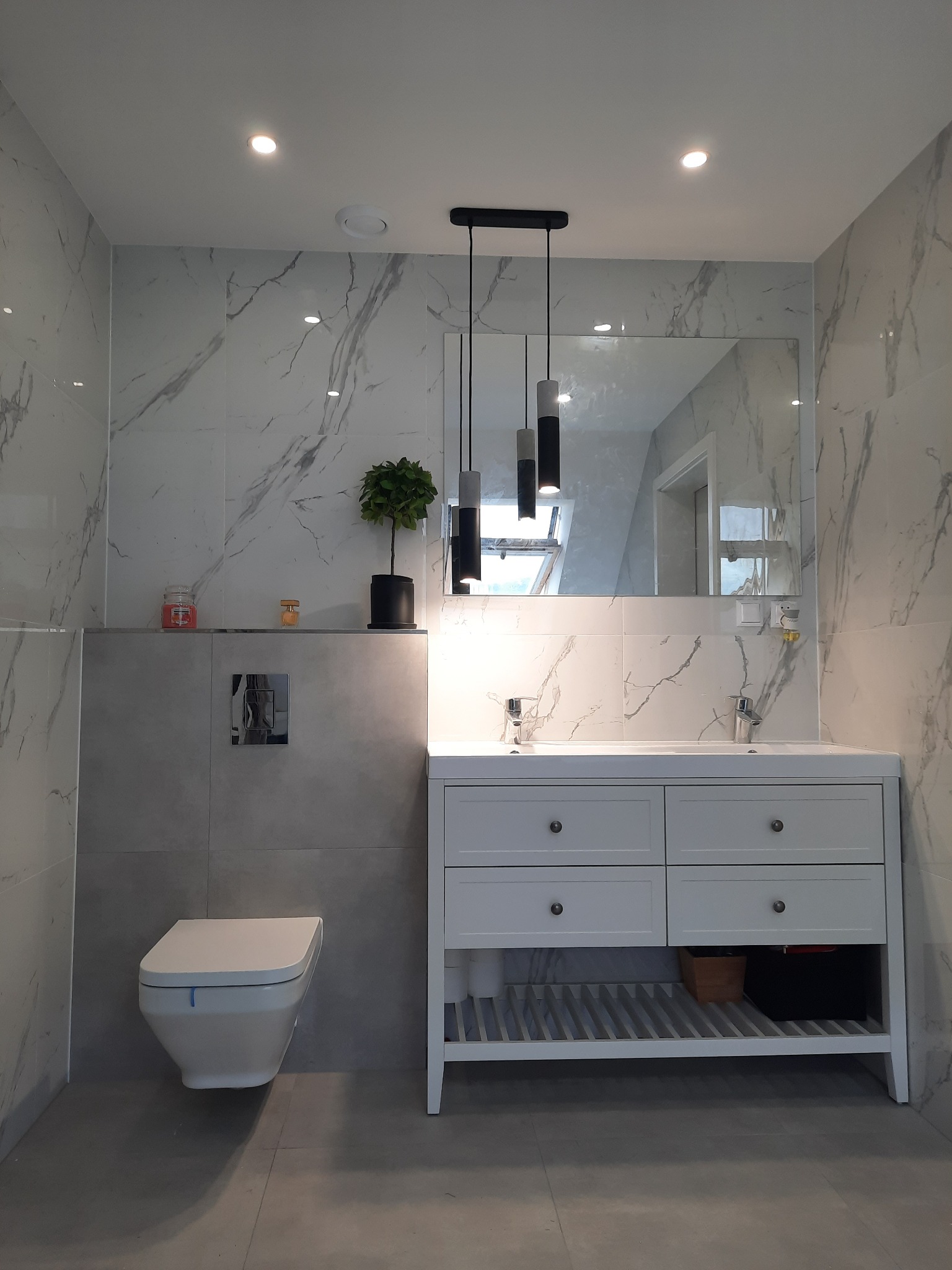 Nowoczesna łazienka z marmurowymi płytkami, wiszącą toaletą, białą szafką pod umywalką i potrójną lampą wiszącą z cylindrycznymi kloszami.