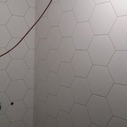 Ameliizlat Team - Ściana łazienki wyłożona białymi, sześciokątnymi płytkami ceramicznymi z widocznym przewodem elektrycznym i przygotowanymi otworami pod armaturę.