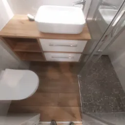 Nowoczesna łazienka po remoncie: biała umywalka nablatowa na drewnianej szafce z szufladami, wisząca toaleta i kabina prysznicowa z mozaiką na podłodze, całość wykończona w jasnych płytkach...