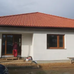 Nowy dom jednorodzinny z białymi ścianami zewnętrznymi i brązową dachówką, widoczne drewniane okna i drzwi, pracownik w czerwonym ubraniu wchodzi do środka, teren wokół domu jeszcze...