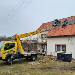 Żółty podnośnik koszowy z numerem telefonu na tle domu z czerwoną dachówką, gdzie dwóch monterów instaluje panele słoneczne na dachu, a na ziemi leżą dodatkowe panele fotowoltaiczne.