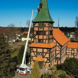Podnośnik koszowy na tle zabytkowego kościoła z zieloną wieżą, podczas prac na wysokości przy szczycie dachu.
