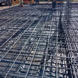 Siatka zbrojeniowa ułożona na czarnej folii, przygotowana do wylania fundamentów, widoczne pionowe pręty i fragmenty osób w tle.