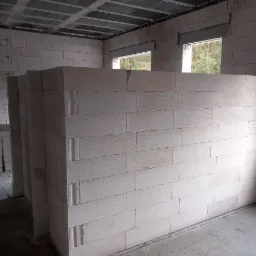 Wnętrze w trakcie budowy z widocznymi ścianami z białych bloczków, surowy betonowy strop i okna z widokiem na drzewa.