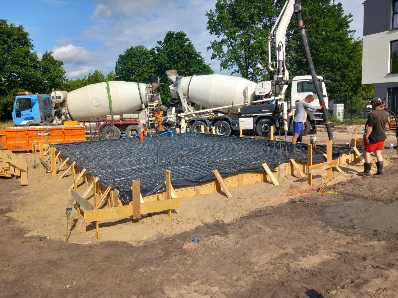 Wylana płyta fundamentowa zbrojona stalową siatką, szalunek drewniany, dwa betoniarki i pracownicy na placu budowy domu jednorodzinnego w słoneczny dzień.