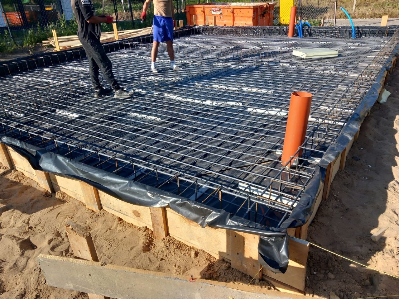 Przygotowanie zbrojenia płyty fundamentowej pod budynek: siatka z prętów stalowych na folii, szalunek z desek, pomarańczowa rura kanalizacyjna wystająca zbrojenia.