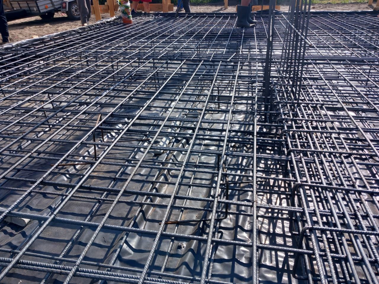 Siatka zbrojeniowa ułożona na czarnej folii, przygotowana do wylania fundamentów, widoczne pionowe pręty i fragmenty osób w tle.