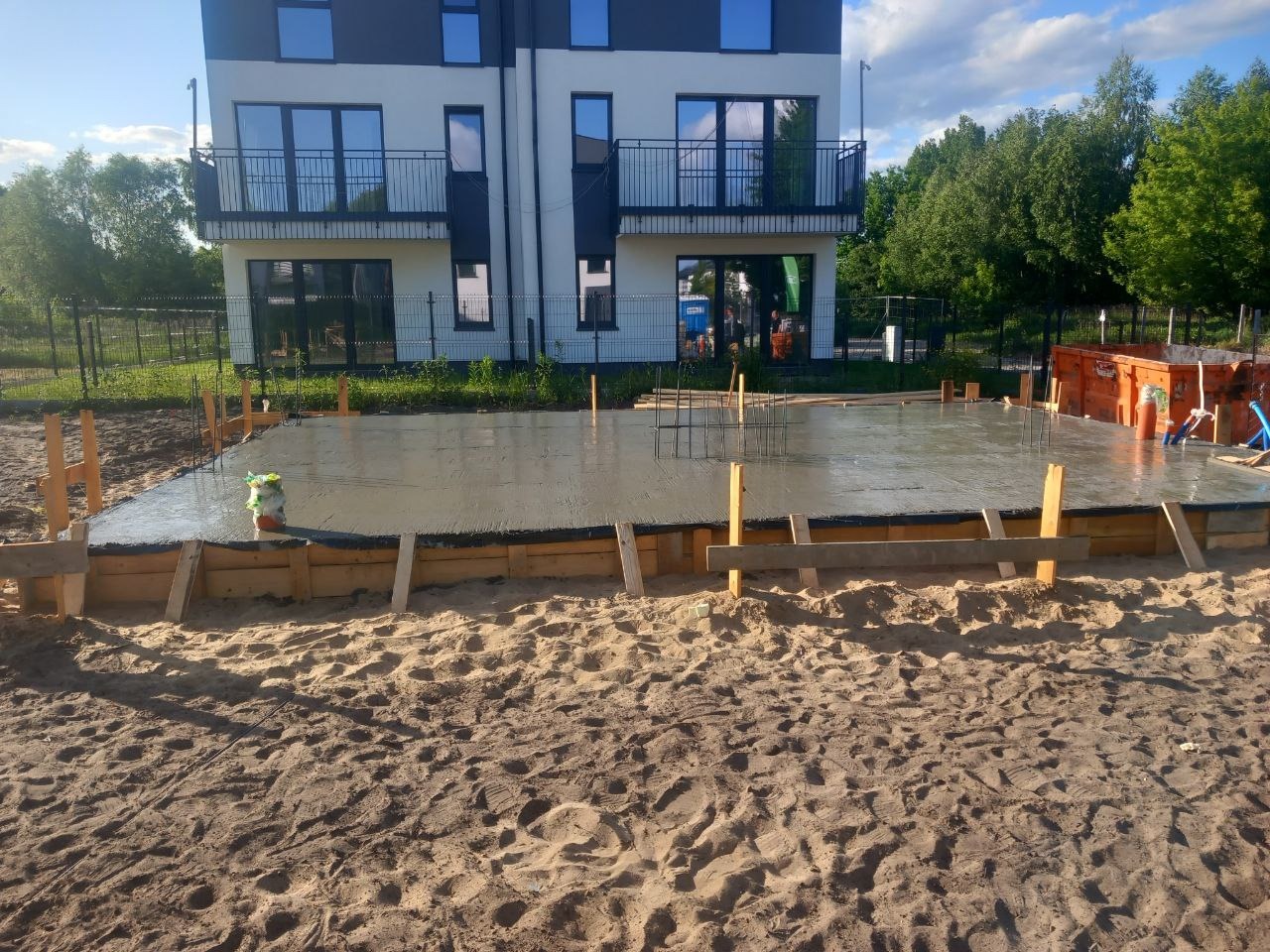Świeżo wylana, mokra płyta fundamentowa z drewnianym szalunkiem na piaszczystym podłożu, widoczne fragmenty zbrojenia i budynek w tle.