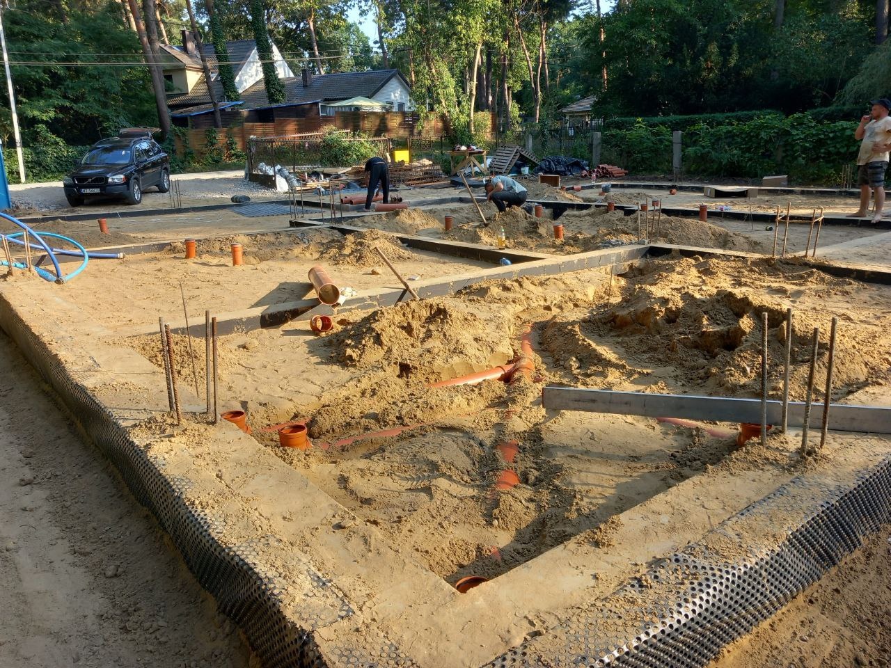 Fundamenty domu w trakcie budowy: widoczne zbrojenia, rury kanalizacyjne w piasku, izolacja pionowa fundamentów z folii kubełkowej, pracownicy w tle.