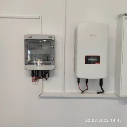 Szara skrzynka bezpiecznikowa i biały falownik Solis S5 zamontowane na białej ścianie, widoczne kable połączeniowe.