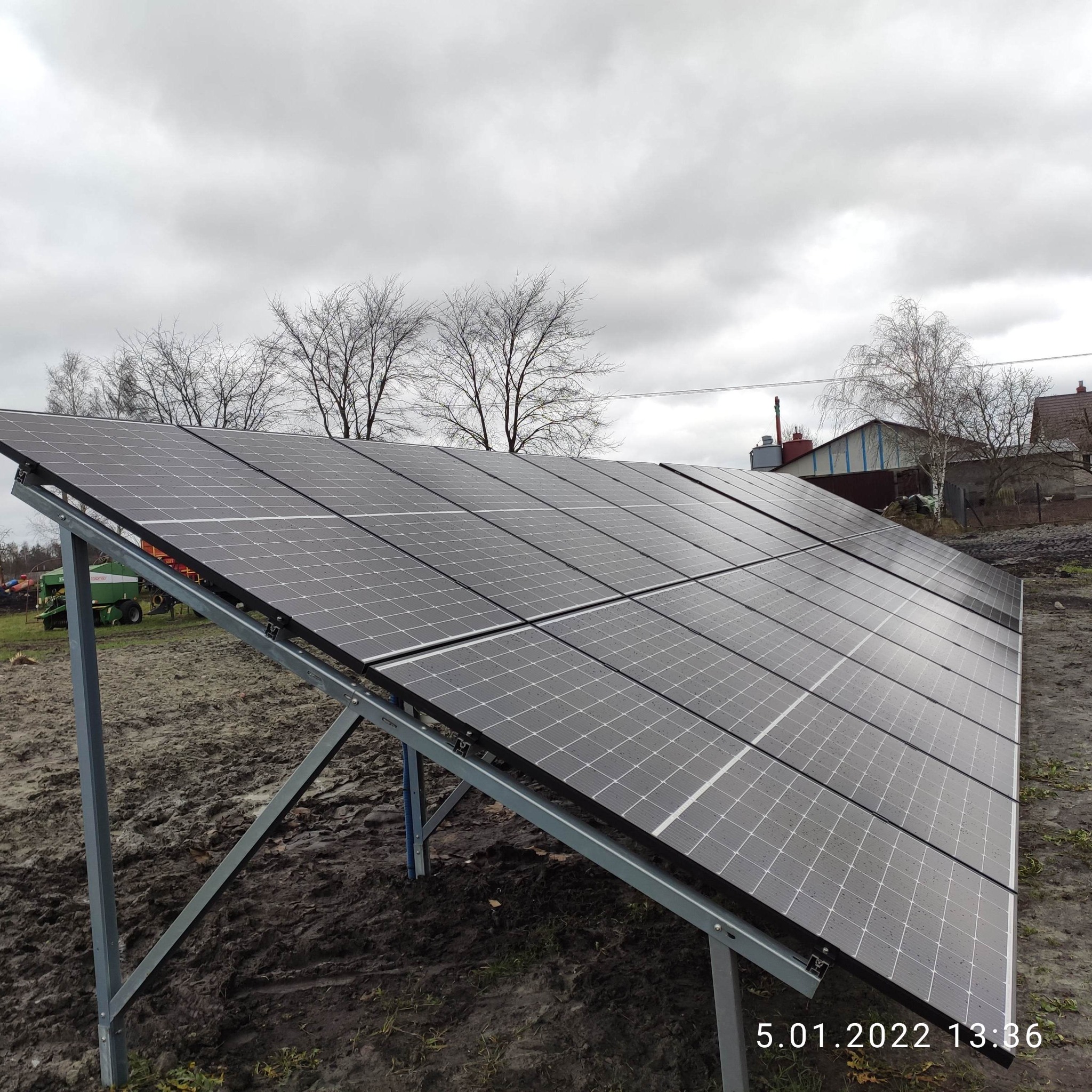 Panel słoneczny zamontowany na metalowej konstrukcji na błotnistym terenie, z widocznym traktorem w tle i pochmurnym niebem.