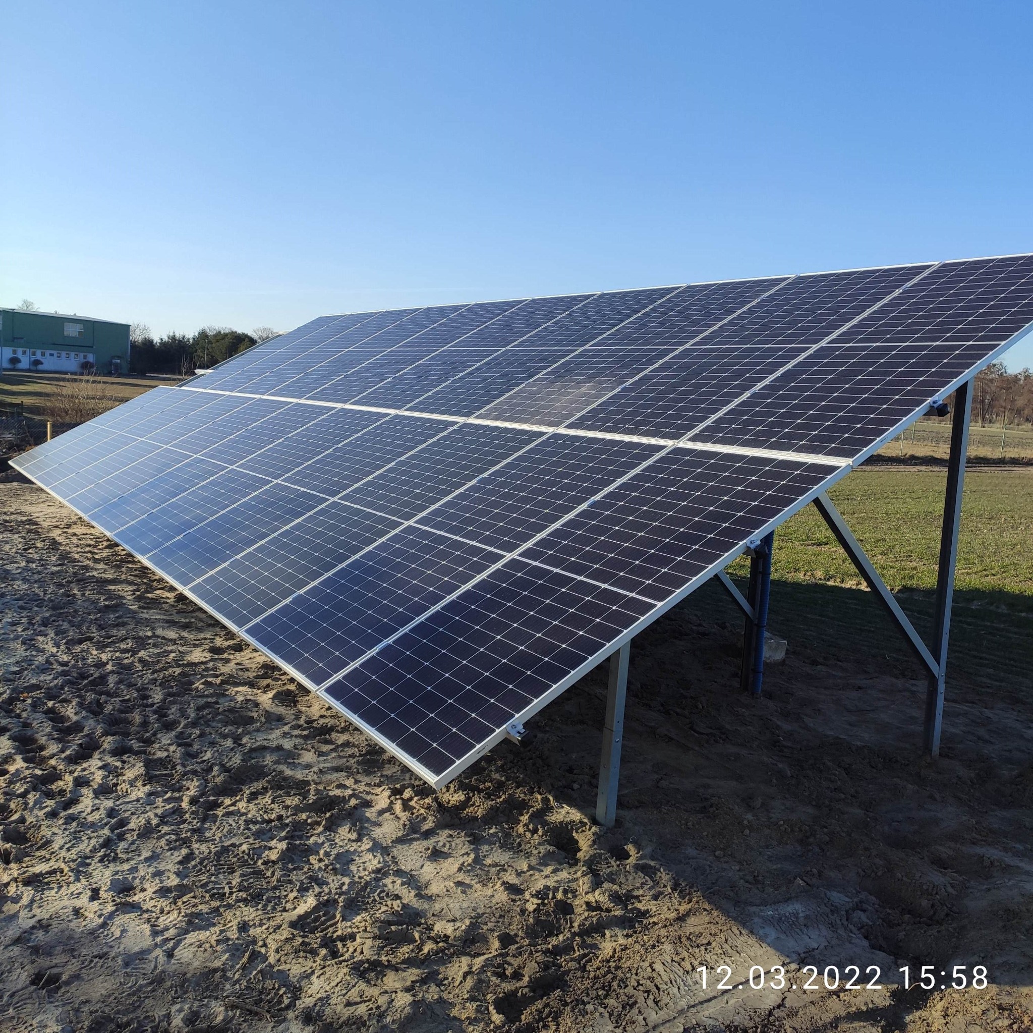 Panel fotowoltaiczny na metalowej konstrukcji, ustawiony na piaszczystym podłożu, w tle budynek i błękitne niebo.