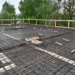 Zbrojenie stropu żelbetowego budynku wielorodzinnego z widoczną siatką zbrojeniową, deskowaniem i wystającymi prętami zbrojeniowymi. Widok z góry na konstrukcję.