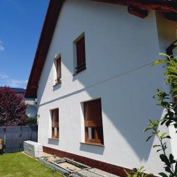 AMH BUD SP. Z O.O. - Biała elewacja domu po naprawie, widoczne brązowe okna z roletami, czerwony dach i fragment rusztowania na trawniku. Słoneczny dzień.