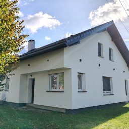 AMH BUD SP. Z O.O. - Jasny, jednorodzinny dom z nową, białą elewacją w słoneczny dzień. Widoczne okna z roletami, ciemny dach i fragment drzewa z żółtymi liśćmi.