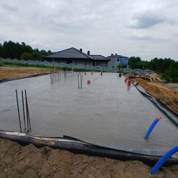 AMH BUD SP. Z O.O. - Wylana płyta fundamentowa z wystającymi prętami zbrojeniowymi i rurami instalacyjnymi. Widok na budynek w tle, pochmurne niebo.
