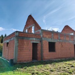 AMH BUD SP. Z O.O. - Wznoszone ściany parteru i poddasza domu jednorodzinnego z czerwonej cegły, z widocznymi betonowymi wzmocnieniami, na tle błękitnego nieba i zieleni.
