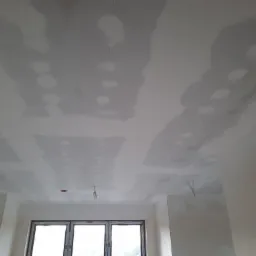 Płyta kartonowo-gipsowa na suficie w trakcie szpachlowania, widoczne łączenia i otwory na oświetlenie, okno w tle.