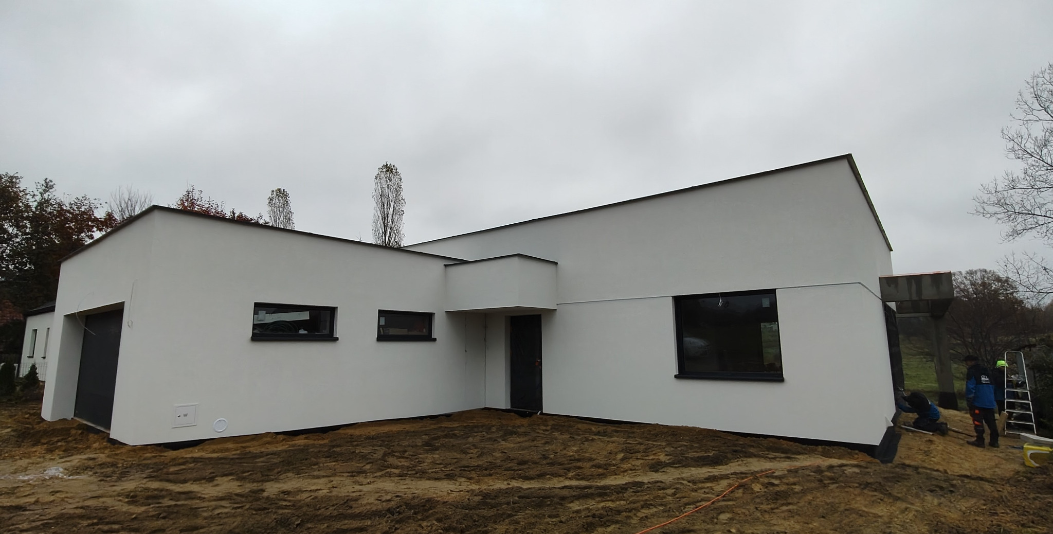 Nowoczesny dom murowany z ocieploną, białą elewacją i czarnymi ramami okiennymi, widoczni pracownicy przy wykończeniu cokołu.