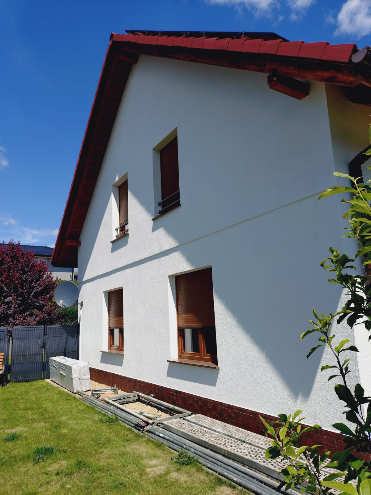 Biała elewacja domu po naprawie, widoczne brązowe okna z roletami, czerwony dach i fragment rusztowania na trawniku. Słoneczny dzień.