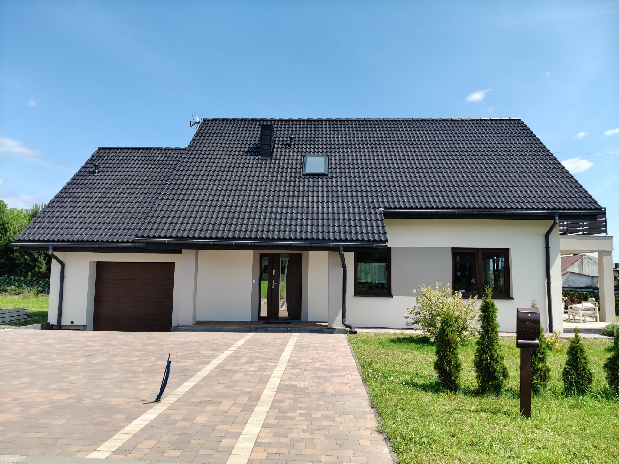 Nowoczesny dom z ocieploną, jasną elewacją i ciemną dachówką. Widoczny brązowy garaż i okna. Kostka brukowa prowadzi do wejścia. Błękitne niebo w tle.