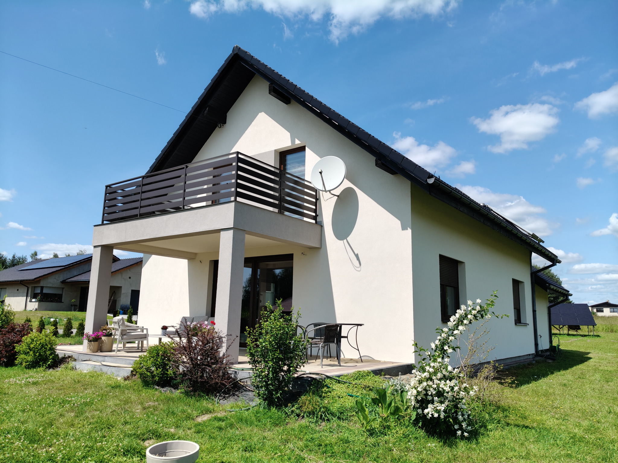 Otynkowany, ocieplony dom jednorodzinny z balkonem i anteną satelitarną w słoneczny dzień. Widoczne elementy wykończenia i zagospodarowania terenu.