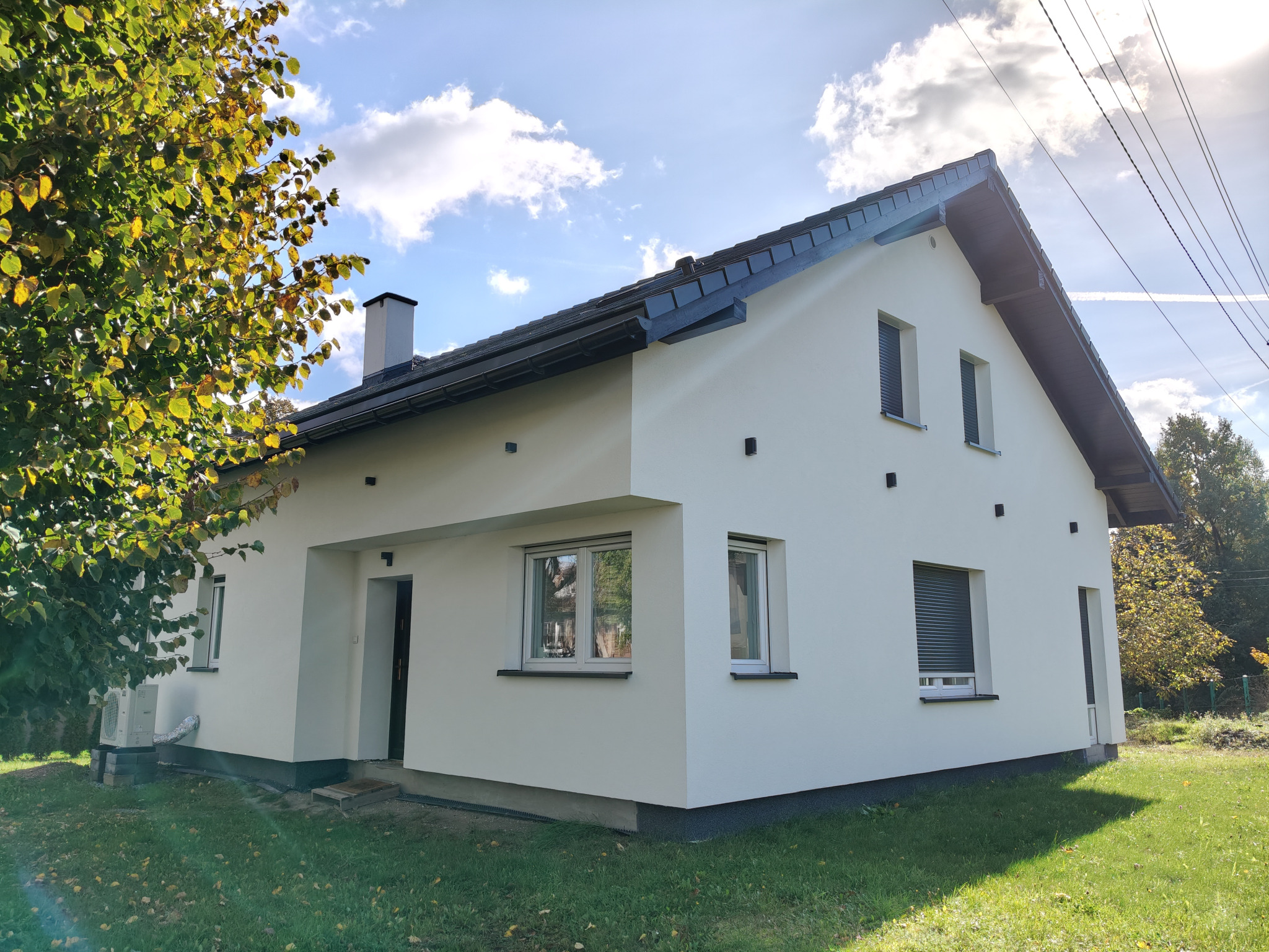 Jasny, jednorodzinny dom z nową, białą elewacją w słoneczny dzień. Widoczne okna z roletami, ciemny dach i fragment drzewa z żółtymi liśćmi.